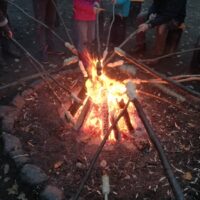 Ein Lagerfeuer brennt und Menschen, im Kreis darum stehend, halten lange Stöcke mit Brotteig darüber.