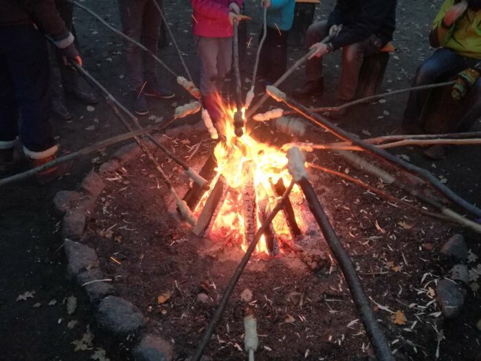 Ein Lagerfeuer brennt und Menschen, im Kreis darum stehend, halten lange Stöcke mit Brotteig darüber.