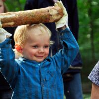 Fröhliches, kleines Kind mit Arbeitshandschuhen hält über dem Kopf einen Holzscheit, um ihn zu werfen.