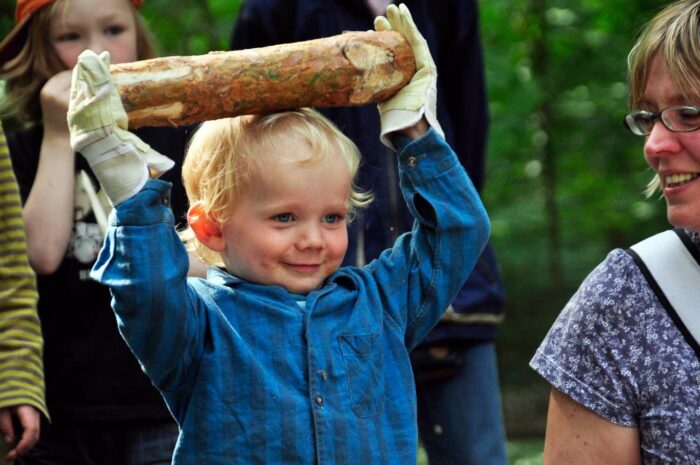 Fröhliches, kleines Kind mit Arbeitshandschuhen hält über dem Kopf einen Holzscheit, um ihn zu werfen.
