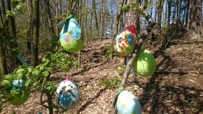 Aus Zeitungsschnipseln, die um Luftballons geklebt wurden, gebastelte Ostereier, bunt bemalt, hängen an Zweigen im Wald.