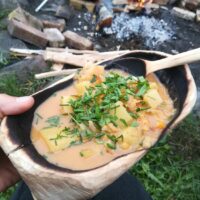 Suppe mit Kräutern in handgefertigtem Holzgeschirr vor Lagerfeuer