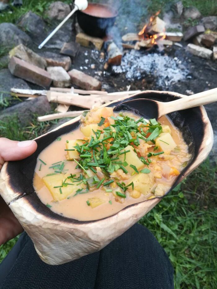 Suppe mit Kräutern in handgefertigtem Holzgeschirr vor Lagerfeuer