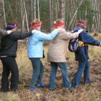 Menschen mit verbundenen Augen, Hand auf der Schulter des/der Vorlaufenden, gehen in einer Reihe durch den Wald.