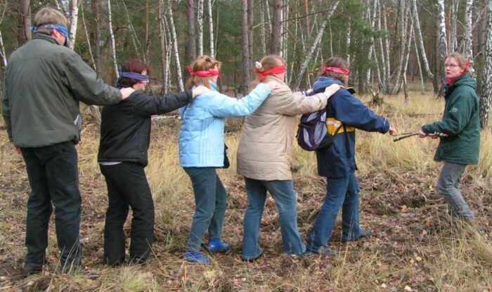 Menschen mit verbundenen Augen, Hand auf der Schulter des/der Vorlaufenden, gehen in einer Reihe durch den Wald.