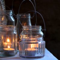 Teelichter brennen in farblosen Gläsern mit Drahthenkel, stehen in der Dunkelheit auf einem Tisch.