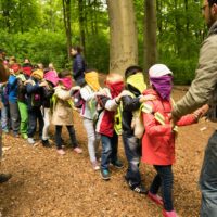 Kinder mit verbundenen Augen, Hand auf der Schulter des/der Vorlaufenden, tasten sich in einer Reihe durch den Wald.